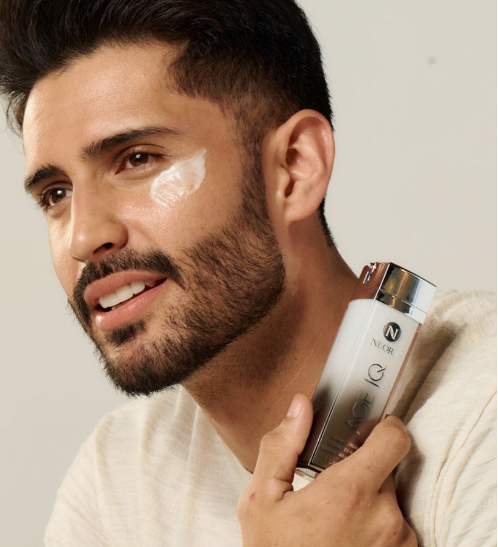 A man with face cream on his under-eye holds up a bottle of Age IQ Night Cream to his neck and shoulder.